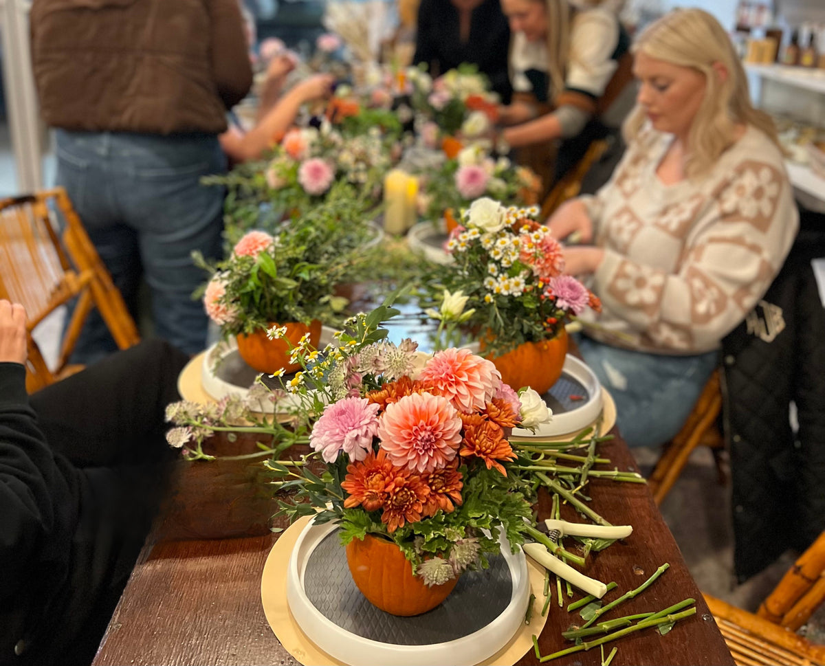 A long table filled with floral arrangements in orange pumpkins, surrounded by people in a warmly lit indoor event space.