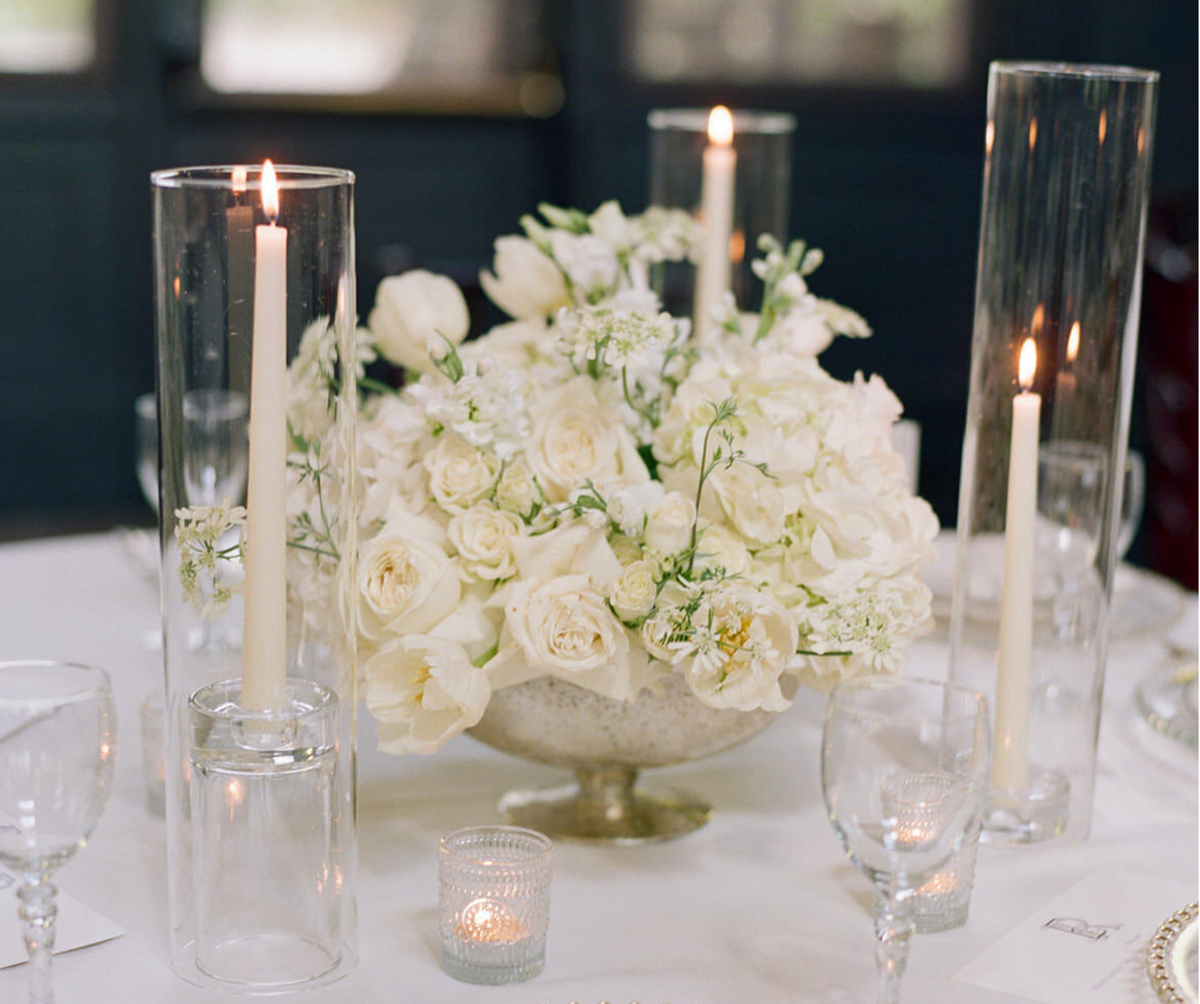 A white floral centerpiece arranged in a silver bowl on a white table, surrounded by tall clear glass candle holders with lit taper candles, wine glasses, and small votive candles in a softly blurred indoor setting.